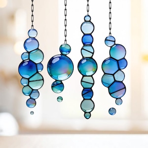 Op de afbeelding: Vier hangende glas-in-lood ornamenten in blauwe en teal tinten, die op bellen lijken. Elk ornament is opgehangen aan een zwarte ketting. De glazen stukken zijn in verschillende maten en vormen, met een iriserende afwerking.