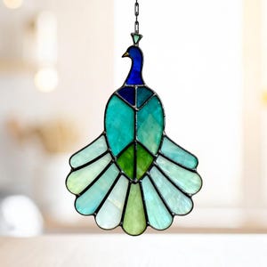 May include: Stained glass peacock suncatcher with a chain for hanging. The peacock features shades of blue, teal, and green, with a black outline. The design includes a crown and a fan-shaped tail, perfect for window decor.