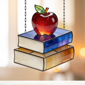May include: A stained glass suncatcher featuring a red apple atop two stacked books. The books have blue and yellow pages with white edges, and the apple has a green leaf. The suncatcher hangs from a black chain.