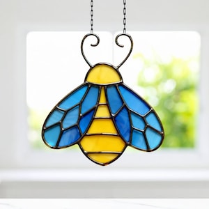 Puede incluir: Atrapasoles de abeja de vidriera con paneles amarillos y azules, contorneados en negro. La abeja está suspendida por una cadena de metal. Las alas son un mosaico de vidrio azul, y el cuerpo es amarillo. Artículo decorativo.