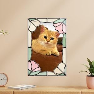 May include: A framed photograph of a golden-colored kitten with big eyes, resting on a brown surface. The frame is stained glass with floral designs in pink, green, and cream colors.