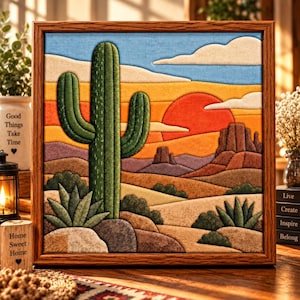 May include: A framed fabric wall art piece depicting a desert landscape with a large green saguaro cactus, smaller agave plants, and rocky formations under a bright red sun. The scene features layered textures in shades of orange, yellow, and blue.