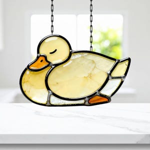 May include: A stained glass duck suncatcher with a yellow body, orange beak, and black outline. The duck is hanging from a chain. The duck is in a sleeping position.