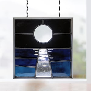 Könnte beinhalten: Glasfenster mit einer Nachtszene mit Vollmond und einem Lichtweg über dem Wasser. Das Panel hat eine quadratische Form mit einem silbernen Metallrahmen und wird von einer schwarzen Kette gehalten. Die Farben sind Schwarz, Blau und Silber.