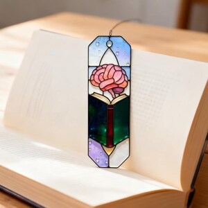 May include: A stained glass bookmark featuring a pink brain over an open book. The bookmark has a blue, green, and purple color scheme with a metal loop at the top. The bookmark is placed in an open book.
