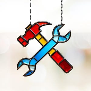 May include: A stained glass decoration featuring a hammer and wrench crossed. The hammer is red with a yellow handle, and the wrench is blue with red and yellow accents. The tools are suspended by black chains.