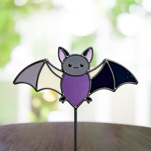 May include: A colorful bat-shaped garden stake with a cheerful expression. The bat features a gray head, purple body, and wings in shades of gray, white, and navy blue. The decorative piece is mounted on a thin, black metal rod.