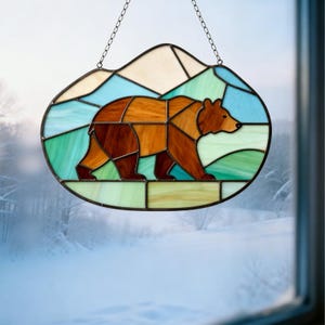 Puede incluir: Vidriera con un oso marrón y naranja, sobre un fondo de formas geométricas azules, verdes y crema. La obra está enmarcada en negro y cuelga de una cadena. El fondo muestra un paisaje nevado.