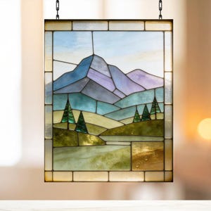 Könnte beinhalten: Buntglasfenster mit einer Berglandschaft mit einer blau-violetten Bergkette, grünen Bäumen und sanften Hügeln. Das Fenster ist in dunklem Metall gerahmt und hängt an einer Kette. Das Glas weist eine Vielzahl von Farben auf.