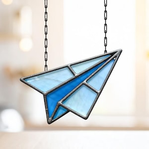 May include: A stained glass paper airplane in shades of blue, suspended by a chain. The design features a dark metal frame and varying tones of blue glass, creating a decorative piece.