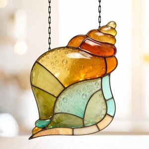 May include: Stained glass seashell suncatcher with a variety of colors including amber, yellow, green, and teal. The seashell is segmented and has a textured appearance. It hangs from a black chain.
