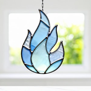 May include: A stained glass flame in shades of blue and white, with a black metal frame and a chain for hanging. The flame design is composed of multiple glass pieces, creating a textured appearance. The background is a blurred view of a window.