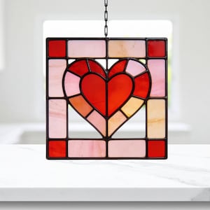 Puede incluir: Un panel de vidriera con un diseño de corazón. El corazón está compuesto por piezas de vidrio rojas y rosas, rodeado por un marco cuadrado con vidrio rojo, rosa y color melocotón. El panel está suspendido por una cadena negra.