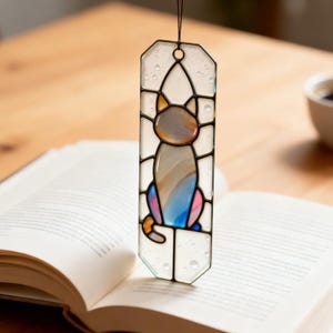 May include: A vertical stained glass suncatcher featuring a stylized cat silhouette. The cat is composed of iridescent, blue, and pink glass segments, set within a clear, textured glass frame with dark metal soldering and a hanging loop.
