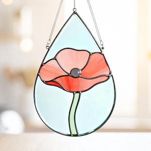 May include: Stained glass poppy flower suncatcher in a teardrop shape. The flower has red and pink petals with a gray center. The stem is green, and the background is light blue. The suncatcher is framed with a dark metal and hangs from a chain.