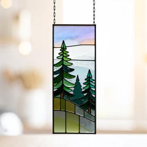 May include: A rectangular stained glass panel featuring a mountain landscape with evergreen trees. The panel has a black metal frame and is suspended by a chain. The trees are various shades of green, and the sky is a mix of blue and pink.