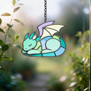 Könnte beinhalten: Ein Glasfenster-Drachen-Suncatcher in Blau-, Grün- und Lilatönen mit schwarzem Metallumriss. Der Drache ist zusammengerollt, die Augen geschlossen, und hängt an einer Kette. Der Hintergrund ist eine verschwommene Ansicht von Grün.