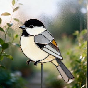 May include: Stained glass bird decoration featuring a chickadee design. The bird has a black head, cream-colored body, and gray and brown wings. It is mounted on a metal rod for display in a garden or as indoor decor.