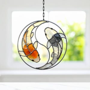 Könnte beinhalten: Glasfenster-Suncatcher mit Koi-Fischen in einem Yin-Yang-Design. Ein Fisch ist orange und weiß, der andere schwarz und weiß. Der Suncatcher wird von einer schwarzen Kette gehalten. Das Design wird von einem kreisförmigen Metallrahmen umrandet.