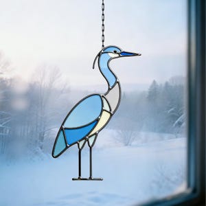 May include: Stained glass heron suncatcher in shades of blue, white, and yellow, with a black metal frame and chain for hanging. The bird is depicted in a stylized, geometric design, perfect for window decor.
