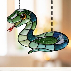 May include: A stained glass snake suncatcher in green and blue hues, with a red tongue and a golden eye. The snake is segmented and suspended by a black chain. Designed to catch the light, it creates a colourful display.