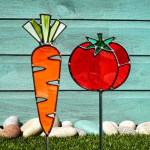 May include: Two stained glass garden stakes: a carrot and a tomato. The carrot is orange with green and white leaves. The tomato is red with green leaves. Both are outlined in dark metal and set in a garden.