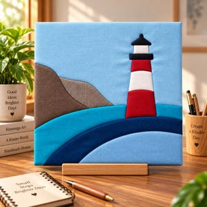 May include: A square fabric wall art piece featuring a lighthouse with red, white, and black sections set against a light blue sky. The scene includes a brown cliffside and layered blue waves in dark and medium tones, all mounted on a wooden stand.