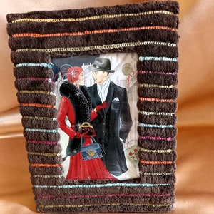 Könnte beinhalten: Ein brauner Bilderrahmen mit einem gestreiften Muster und einem Bild eines Paares in Vintage-Kleidung. Die Frau trägt ein rotes Kleid und einen Pelzmantel, und der Mann trägt einen Anzug und einen Hut.