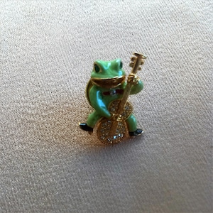 May include: A green frog playing a gold bass guitar with rhinestones. The frog is wearing a gold bow tie.
