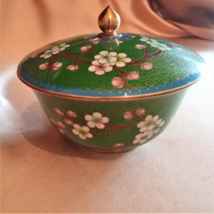 Puede incluir: Un cuenco de cloisonné verde y dorado con tapa. El cuenco está decorado con flores blancas y rosas y detalles dorados. La tapa tiene un pomo dorado.