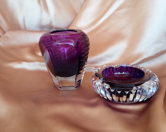 May include: A clear glass vase and ashtray set. The vase is a deep purple color with a faceted base and a textured upper portion. The ashtray is round with a purple interior and a clear, cut-glass exterior. The set is displayed on a satin surface.