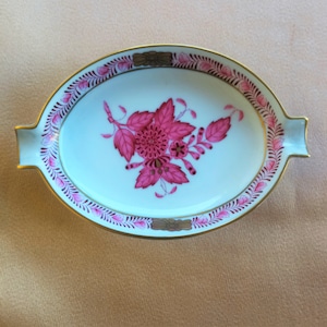 May include: An oval-shaped porcelain ashtray with a pink floral design and a gold rim. The ashtray has a small lip on one side for holding cigarettes.