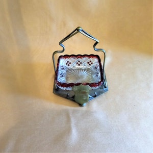 May include: A clear glass dish with a red rim sits in a silver metal stand with a handle. The stand has a green jade-like accent.