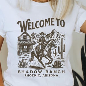 Può includere: T-shirt bianco con un design grafico raffigurante un cowboy a cavallo, una casa, montagne e cactus. Il testo "WELCOME TO SHADOW RANCH PHOENIX, ARIZONA" è stampato sulla maglietta.