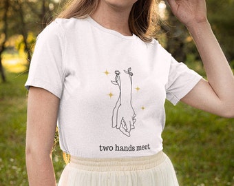 May include: A white short-sleeved t-shirt featuring a black line art graphic of two hands holding each other with small floral accents and gold star symbols. The text "two hands meet" is printed in black lowercase letters below the graphic.