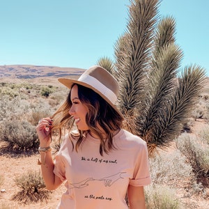 May include: A woman wearing a peach-colored t-shirt with the text "He is half of my soul, as the poets say." The shirt is paired with a floral skirt and a brown hat with a white band. The background features desert vegetation.