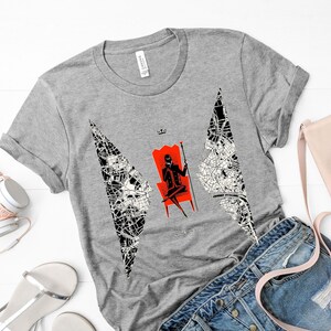Op de afbeelding: Een grijs T-shirt met een zwart-witte afbeelding van een persoon die op een rode troon met een kroon zit. De afbeelding is omringd door een zwart-witte kaart-achtige vormgeving.