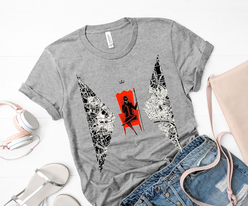Op de afbeelding: Een grijs T-shirt met een zwart-witte afbeelding van een persoon die op een rode troon met een kroon zit. De afbeelding is omringd door een zwart-witte kaart-achtige vormgeving.