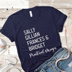Peut inclure: T-shirt bleu marine avec le texte blanc "SALLY GILLIAN FRANCES & BRIDGET Practical Magic". Le t-shirt est pli&eacute;, avec un casque, un sac &agrave; main et un short en jean en arri&egrave;re-plan.