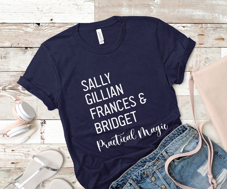 Peut inclure: T-shirt bleu marine avec le texte blanc "SALLY GILLIAN FRANCES & BRIDGET Practical Magic". Le t-shirt est pli&eacute;, avec un casque, un sac &agrave; main et un short en jean en arri&egrave;re-plan.