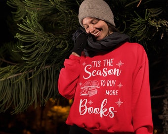 May include: A red sweatshirt with the text "'Tis the Season to Buy More Books" in white. The design includes snowflakes and a stack of books. The person is wearing a gray beanie, black gloves, and a dark gray scarf.