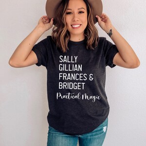 Peut inclure: T-shirt gris fonc&eacute; avec les noms Sally, Gillian, Frances & Bridget et les mots "Practical Magic" en &eacute;criture blanche. Le t-shirt est port&eacute; avec un jean bleu et un chapeau marron.