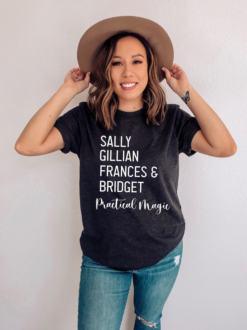 Peut inclure: T-shirt gris fonc&eacute; avec les noms Sally, Gillian, Frances & Bridget et les mots "Practical Magic" en &eacute;criture blanche. Le t-shirt est port&eacute; avec un jean bleu et un chapeau marron.