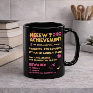 May include: Black ceramic mug with the text "NEEEW ACHIEVEMENT" in pink and yellow. The mug also features text about pregnancy milestones, including "Progress: 12% Complete" and "Reward: +5 Appetite".