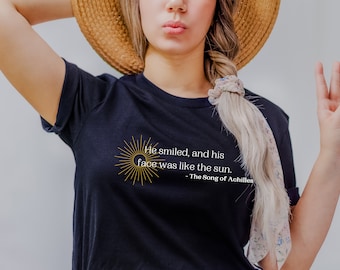 May include: A dark navy blue short-sleeved t-shirt featuring a gold sun graphic and white text that reads: He smiled, and his face was like the sun. - The Song of Achilles. The shirt has a crew neckline and a standard fit for casual wear.