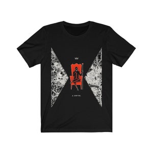Op de afbeelding: Zwart T-shirt met een witte afbeelding van een persoon die op een rode troon zit met een kroon en een scepter. De afbeelding is omgeven door een wit kaartontwerp. De tekst "A NOVEL" staat onder de afbeelding.