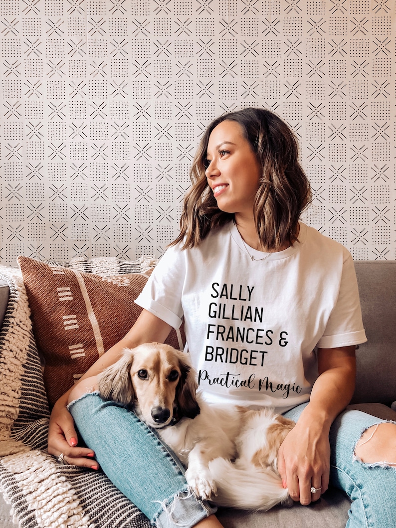 Peut inclure: Un t-shirt blanc avec le texte "SALLY GILLIAN FRANCES & BRIDGET Practical Magic" en noir. Une femme porte le t-shirt et est assise sur un canap&eacute; avec un chien. Le chien est brun clair et blanc.