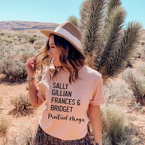 Peut inclure: T-shirt rose clair avec l'inscription noire "SALLY GILLIAN FRANCES & BRIDGET Practical Magic". La personne porte un chapeau marron avec un ruban blanc et une jupe &agrave; imprim&eacute; floral. L'arri&egrave;re-plan est un paysage d&eacute;sertique.