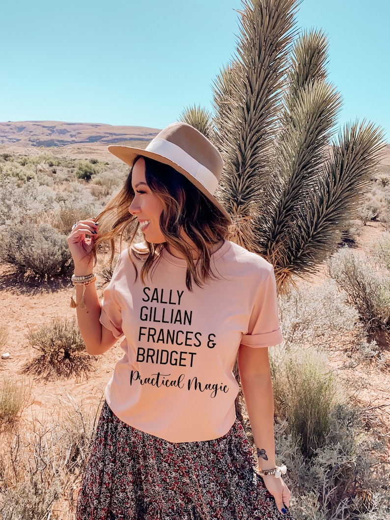 Peut inclure: T-shirt rose clair avec l'inscription noire "SALLY GILLIAN FRANCES & BRIDGET Practical Magic". La personne porte un chapeau marron avec un ruban blanc et une jupe &agrave; imprim&eacute; floral. L'arri&egrave;re-plan est un paysage d&eacute;sertique.