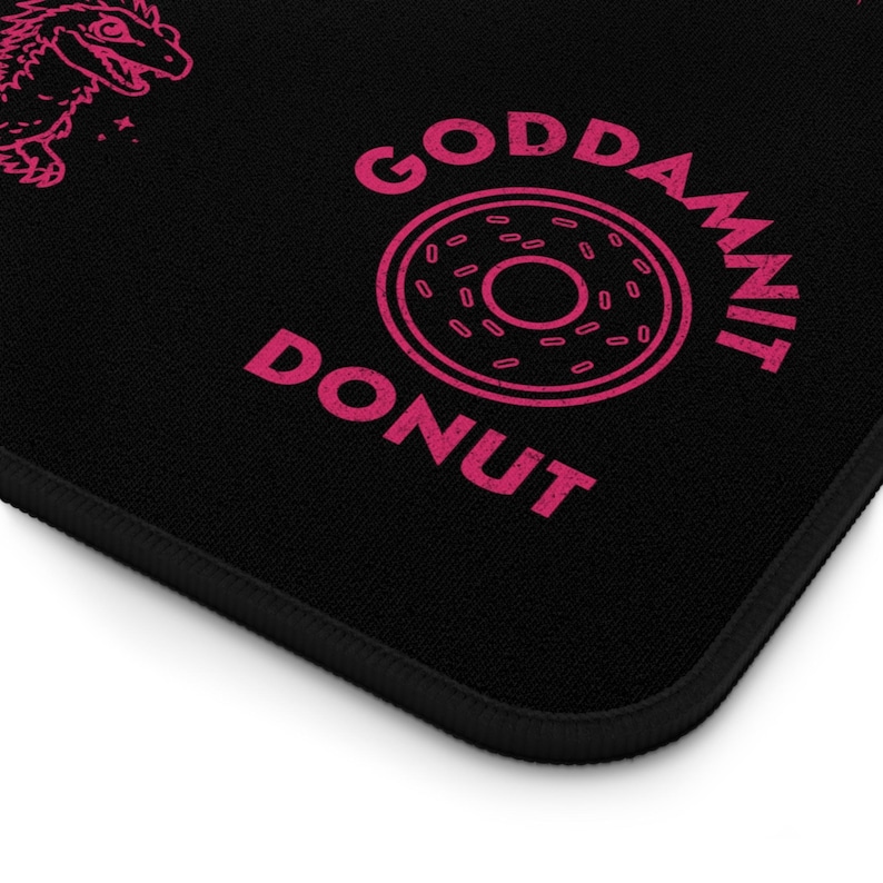 K&ouml;nnte beinhalten: Schwarzes Mousepad mit einem rosa Donut-Motiv und den Worten "GODDAMNIT DONUT". Das Mousepad hat auch ein rosa Dinosaurier-Motiv in der oberen linken Ecke. Die R&auml;nder sind schwarz.